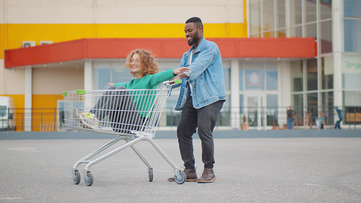 A man in a parking lot is pushing a woman who is sitting in a shopping cart with her hands extended