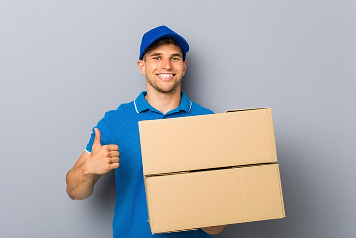Man wearing a blue baseball cap and matching blue polo is giving the thumbs up with one hand while holding a cardboard box with the other