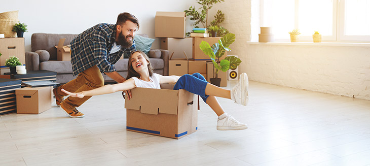 A woman sitting in a cardboard box with her arms extended and legs out is being pushed across the floor by a man as they unpack their belongings in their new home